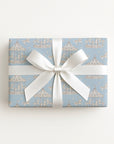 A rectangular gift box wrapped in light blue wrapping paper with a repeating carousel pattern, tied with a wide white satin ribbon and bow, photographed from above on a plain white background.