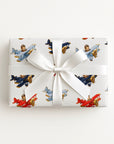A square gift box wrapped in white paper with a repeating pattern of vintage-style airplanes in red, blue, and brown, tied with a white satin ribbon and bow, photographed from above on a light neutral background.