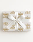 A rectangular gift box wrapped in white paper with a repeating gold carousel pattern, tied with a wide white satin ribbon and bow, photographed from above on a plain white background.