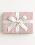 A rectangular gift box wrapped in dusty pink paper with a white line-art pattern and tied with a wide white satin ribbon bow, shown on a clean white background.