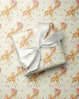 A wrapped gift box with an ivory satin ribbon sits on top of wrapping paper printed with a repeating pattern of babies and giraffes wearing pink hats and bows on a light yellow background.