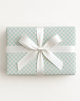 A rectangular gift box wrapped in aquamarine and off white checkered paper with a wide white satin ribbon tied in a bow, photographed from above on a clean white background.
