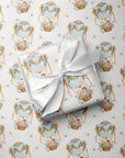 A wrapped baby boy gift box tied with a white satin ribbon, placed on top of matching wrapping paper printed with a repeating vintage hot air balloon motif in soft blue, gold, and pink on a white background.