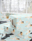 Light aqua wrapping paper with a darling girl ballet dancer pattern, shown wrapped around gift boxes on a table near a window, with scissors and ribbon visible.
