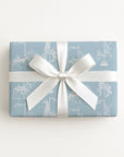 A rectangular gift box wrapped in light blue wrapping paper with a white carousel pattern, tied with a wide white ribbon and bow, on a clean white background.