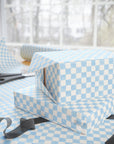 Light blue and white checkered wrapping paper wrapped around gift boxes on a table, with scissors and ribbon visible in a softly lit indoor setting.
