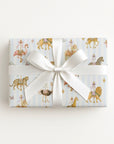 A rectangular gift box wrapped in light blue wrapping paper with a carousel and animal print, tied with a wide white satin ribbon and bow, photographed from above on a clean white background.