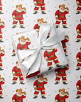 A gift box wrapped in white paper with a large white ribbon bow, placed on top of Christmas-themed wrapping paper featuring a repeating pattern of western Santa Claus in a cowboy outfit.