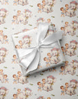 A gift box wrapped in ivory paper with a repeating pattern of vintage-style elephants and children, tied with a white satin ribbon, placed on top of the same patterned wrapping paper.
