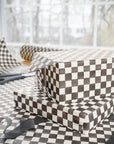 A gift box wrapped in brown and white checkered wrapping paper, with a matching sheet and ribbon on a table in front of a softly blurred window.
