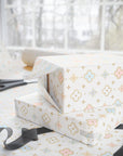 White wrapping paper with a pastel floral and geometric pattern, shown wrapped around gift boxes on a table with scissors and ribbon near a window.