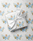 Wrapped gift tied with a white satin bow, displayed on matching wrapping paper featuring a little girl flying a powder blue biplane over a delicate navigation map with gold accents on an ivory background.