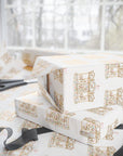 Ivory wrapping paper with a repeating gold carousel illustration pattern, shown wrapped around stacked gift boxes on a table near scissors and ribbon, with a softly blurred window background.