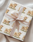 Baby gift wrapped in paper with highland cow and baby pattern and pink ribbon on a satin surface.