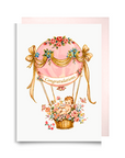 New baby girl card with a baby in a pink floral hot air balloon, golden ribbons and a Congratulations banner
