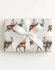 A rectangular gift box wrapped in ivory wrapping paper with a vintage baby and reindeer print, tied with a large white satin ribbon bow, on a clean white background.
