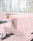 Pink and white checkered wrapping paper displayed on a table with a gift box, scissors, and ribbon in a softly lit room.