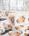 Three gift boxes wrapped in cream baby shower wrapping paper with a pink and gold vintage hot air balloon pattern, styled on a table with scissors and ribbon in front of a softly blurred window.
