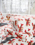 Christmas-themed wrapping paper with a red airplane pattern featuring Santa Claus, shown wrapped around gift boxes on a table with scissors and ribbon near a window.