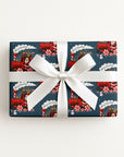 A rectangular gift box wrapped in navy holiday-themed wrapping paper with a red Santa Claus train pattern, tied with a wide white satin ribbon and bow.