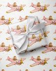Wrapped gift tied with a white satin bow, displayed on matching wrapping paper featuring a little girl flying a pink warplane with a painted shark mouth over a delicate navigation map with gold accents on an ivory background.