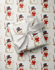 A gift box wrapped in ivory paper with a large white satin bow, placed on top of wrapping paper printed with a repeating Christmas snowman pattern.