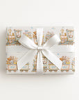A rectangular gift box wrapped in ivory wrapping paper with a whimsical stork and train illustration pattern and tied with a wide white satin ribbon bow.