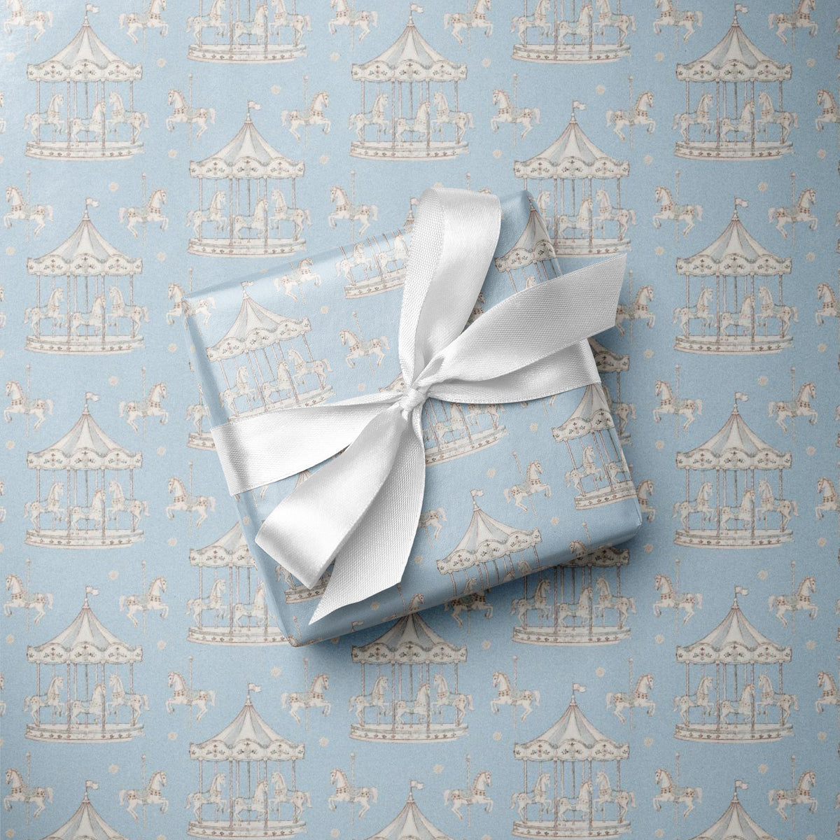A square gift box wrapped in light blue wrapping paper with a repeating carousel pattern, tied with a white satin ribbon, placed on top of the same patterned paper.
