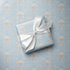 A square gift box wrapped in light blue wrapping paper with a repeating carousel pattern, tied with a white satin ribbon, placed on top of the same patterned paper.