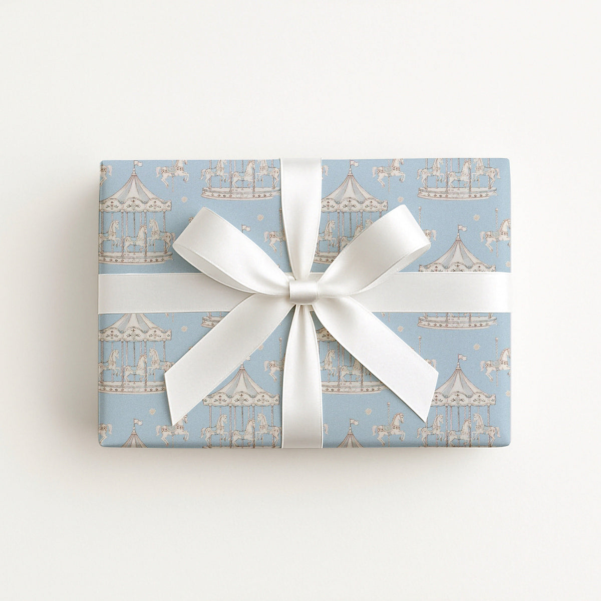 A rectangular gift box wrapped in light blue wrapping paper with a repeating carousel pattern, tied with a wide white satin ribbon and bow, photographed from above on a plain white background.