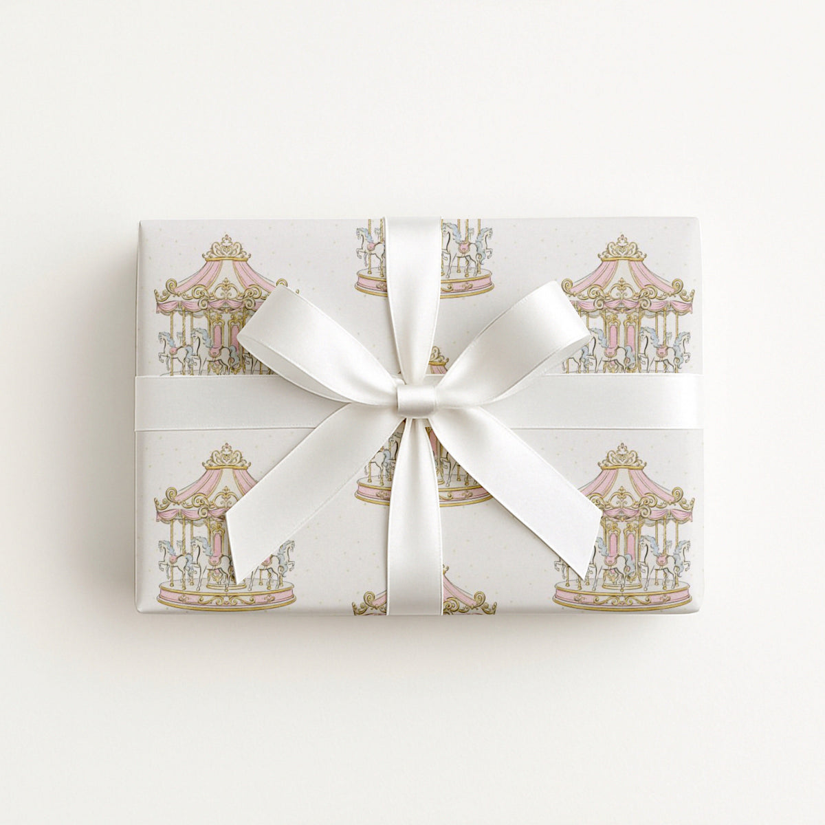 A rectangular gift box wrapped in white paper with a delicate pastel carousel pattern and tied with a wide white satin ribbon bow, photographed against a clean white background.