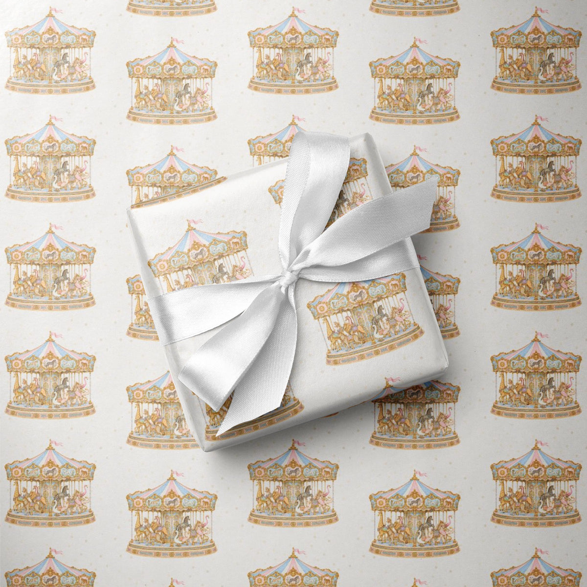 A gift box wrapped in white paper with a large white satin bow, placed on top of wrapping paper printed with a repeating carousel motif in soft gold, beige, and light blue tones.
