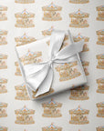 A gift box wrapped in white paper with a large white satin bow, placed on top of wrapping paper printed with a repeating carousel motif in soft gold, beige, and light blue tones.