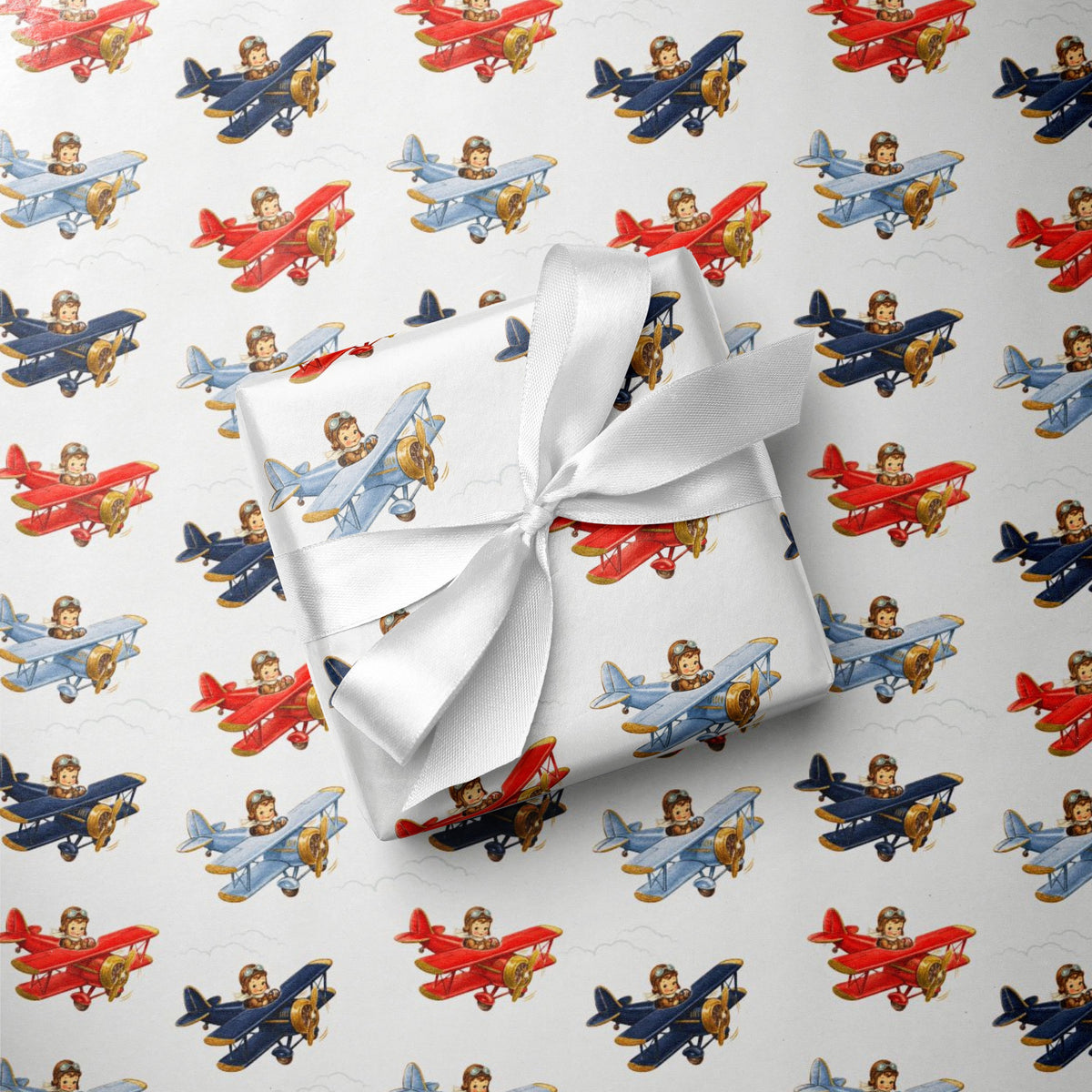 A wrapped gift box tied with a white satin ribbon, placed on top of wrapping paper printed with a repeating pattern of vintage red, blue, and gold biplanes on a white background.