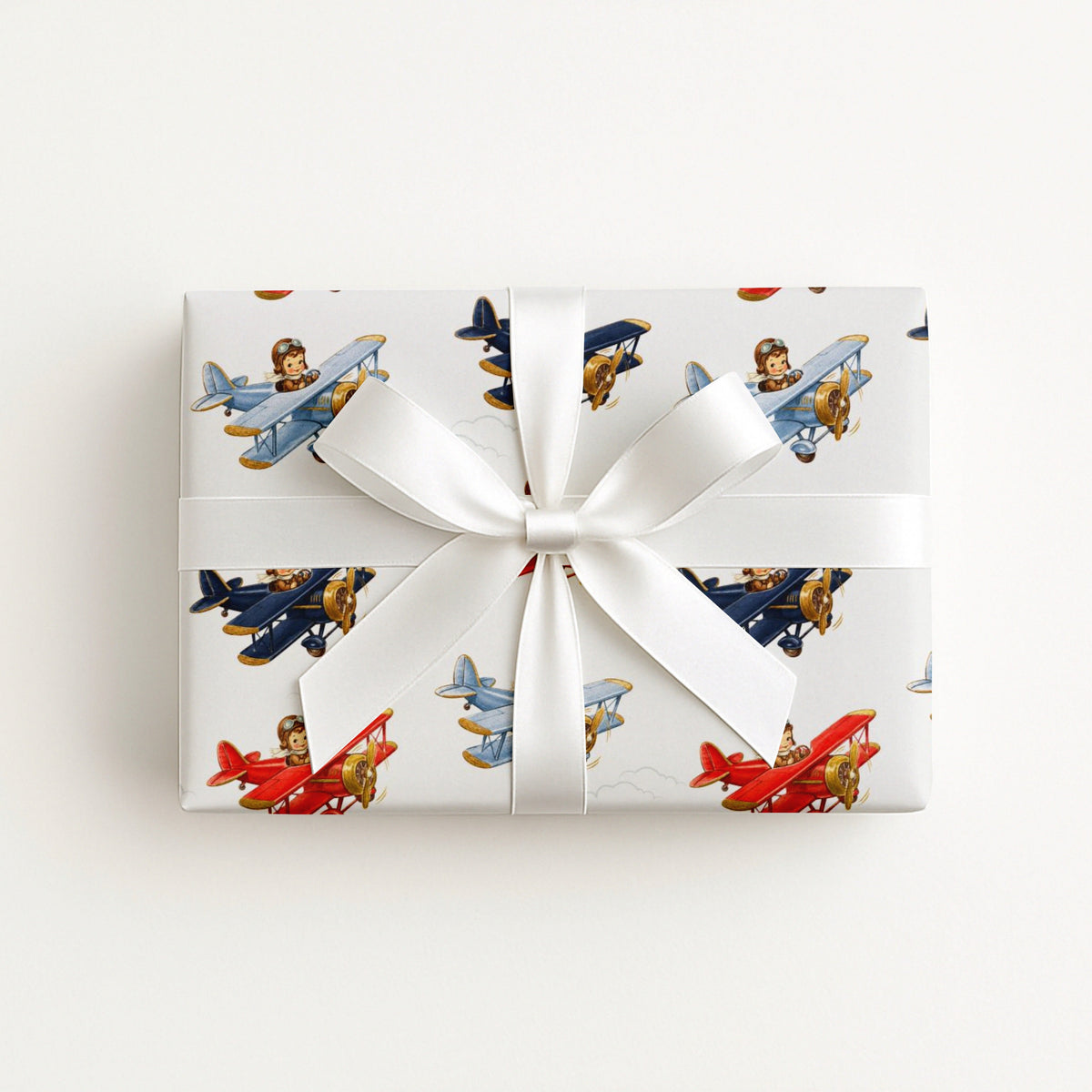 A square gift box wrapped in white paper with a repeating pattern of vintage-style airplanes in red, blue, and brown, tied with a white satin ribbon and bow, photographed from above on a light neutral background.