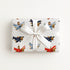 A square gift box wrapped in white paper with a repeating pattern of vintage-style airplanes in red, blue, and brown, tied with a white satin ribbon and bow, photographed from above on a light neutral background.