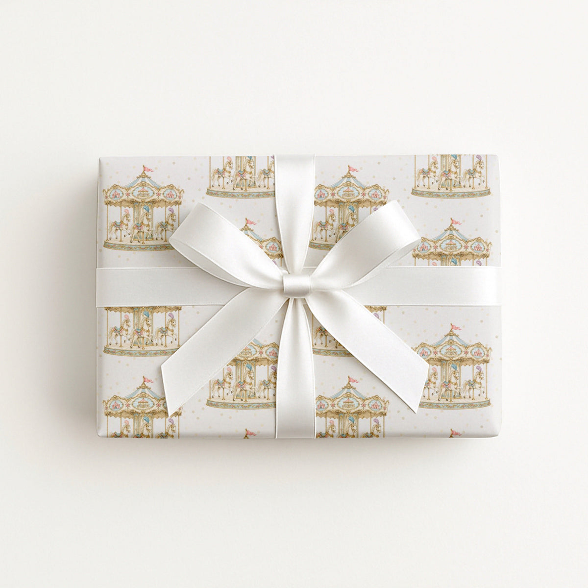 A rectangular gift box wrapped in white paper with a repeating gold carousel pattern, tied with a wide white satin ribbon and bow, photographed from above on a plain white background.