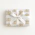 A rectangular gift box wrapped in white paper with a repeating gold carousel pattern, tied with a wide white satin ribbon and bow, photographed from above on a plain white background.