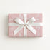A rectangular gift box wrapped in dusty pink paper with a white line-art pattern and tied with a wide white satin ribbon bow, shown on a clean white background.