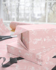 Pink wrapping paper with a white carousel horse pattern, shown wrapped around two gift boxes on a table with scissors and ribbon near a window.