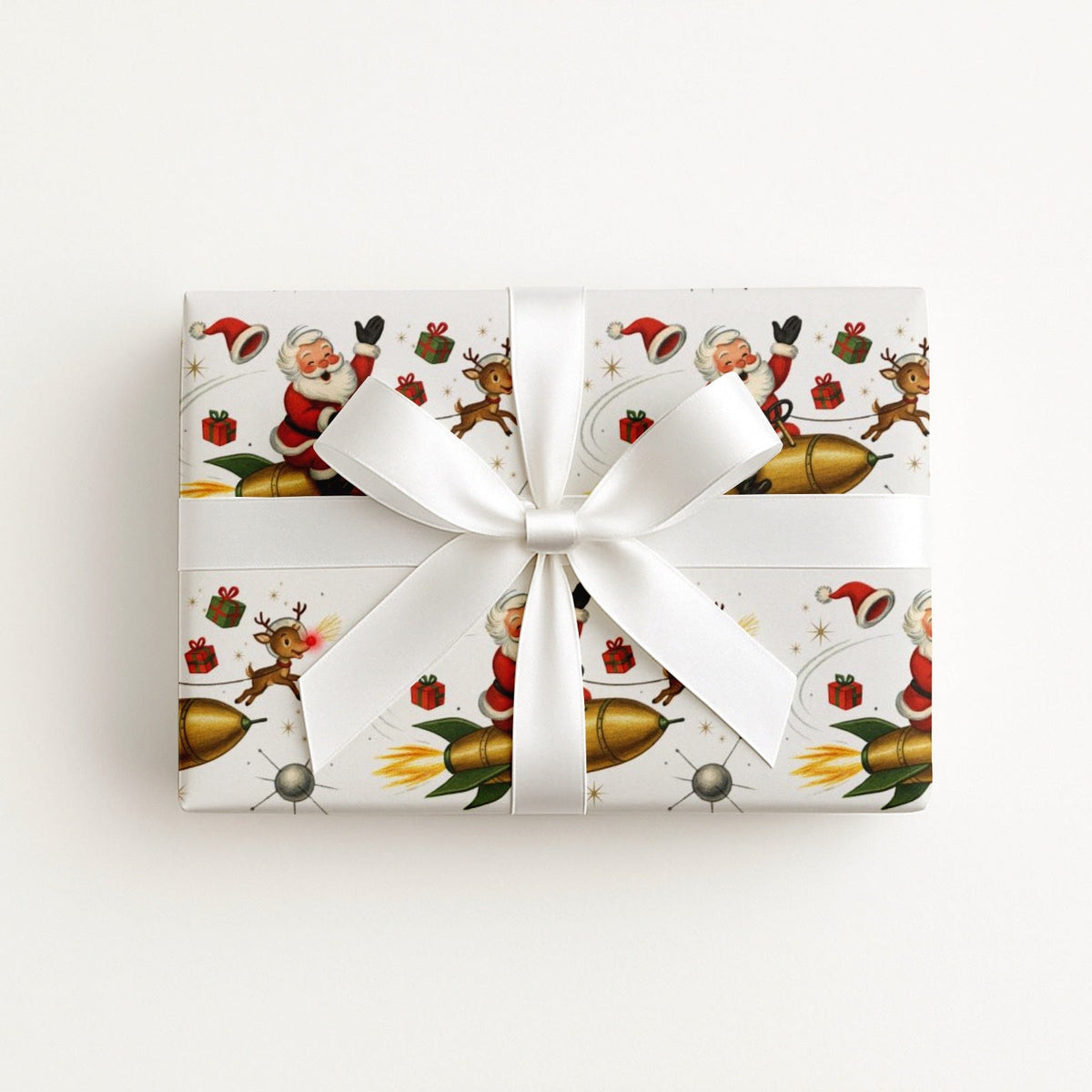 A rectangular gift box wrapped in ivory Christmas-themed paper with Santa Claus, reindeer, ornaments, and gold rockets, tied with a large white satin ribbon and bow.