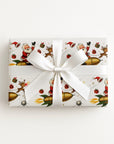 A rectangular gift box wrapped in ivory Christmas-themed paper with Santa Claus, reindeer, ornaments, and gold rockets, tied with a large white satin ribbon and bow.