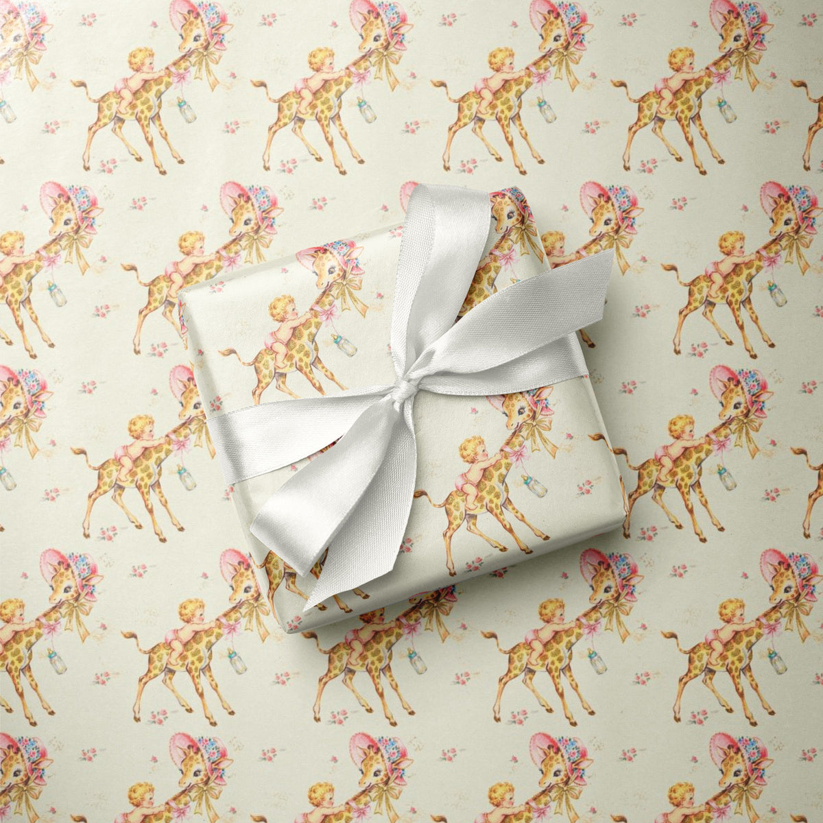 A wrapped gift box with an ivory satin ribbon sits on top of wrapping paper printed with a repeating pattern of babies and giraffes wearing pink hats and bows on a light yellow background.