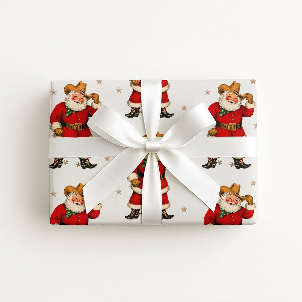 A rectangular gift box wrapped in white paper featuring a repeating print of vintage Santa Claus as a cowboy, tied with a wide white satin ribbon and bow, photographed from above on a plain white background.