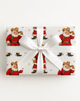 A rectangular gift box wrapped in white paper featuring a repeating print of vintage Santa Claus as a cowboy, tied with a wide white satin ribbon and bow, photographed from above on a plain white background.