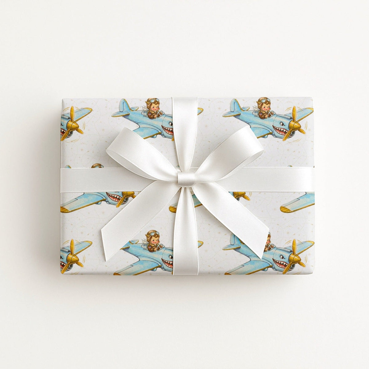 A rectangular gift box wrapped in ivory wrapping paper with a repeating pattern of vintage blue airplanes with shark mouth noses with gold accents, tied with a wide white satin ribbon and bow, on a plain white background.