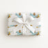 A rectangular gift box wrapped in ivory wrapping paper with a repeating pattern of vintage blue airplanes with shark mouth noses with gold accents, tied with a wide white satin ribbon and bow, on a plain white background.