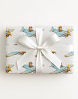 A rectangular gift box wrapped in ivory wrapping paper with a repeating pattern of vintage blue airplanes with shark mouth noses with gold accents, tied with a wide white satin ribbon and bow, on a plain white background.