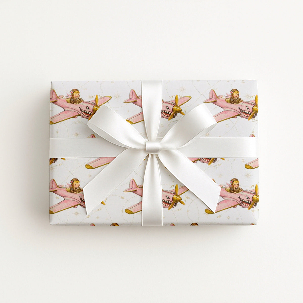 A rectangular gift box wrapped in ivory wrapping paper with a repeating pattern of vintage pink airplanes with shark mouth noses and gold accents, tied with a wide white satin ribbon and bow, on a plain white background.