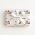 A rectangular gift box wrapped in ivory wrapping paper with a repeating pattern of vintage pink airplanes with shark mouth noses and gold accents, tied with a wide white satin ribbon and bow, on a plain white background.